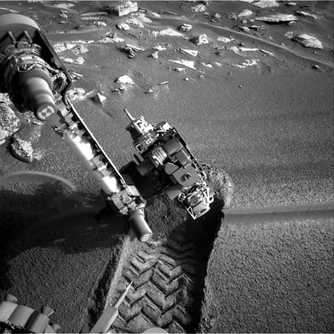 A black-and-white image taken on the surface of Mars. From the upper left, the Curiosity rover's mechanical arm extends downward, holding a large, complex cluster of scientific instruments (the turret) close to the ground. Directly beneath the turret is a deep, freshly made rover wheel track featuring a distinct zig-zag tread pattern pressed into the fine, dark soil. In the background, the sandy terrain transitions to scattered, flat, light-colored rocks. Sharp shadows indicate bright, direct su