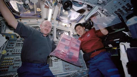 two male astronauts hold up a bag full of small american flags inside a spacecraft