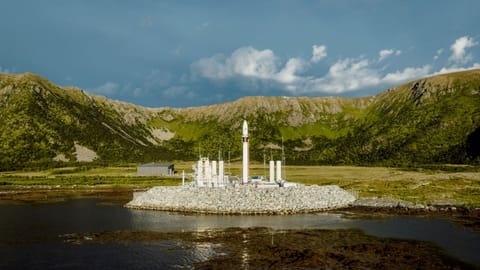 a white rocket stands on a seaside pad with green mountains in the background