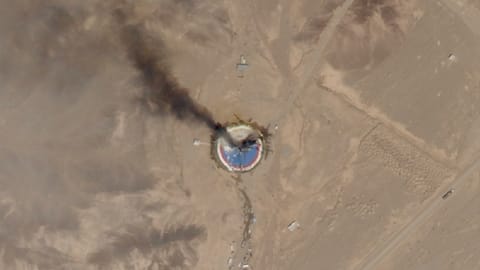 satellite photo of a dark plume of smoke ascending from a blue and red circular launch pad in a desert landscape