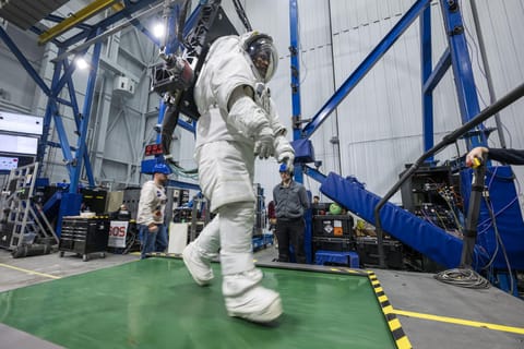 Dressed in a white space suit—with hands gloved, feet booted and head in a helmet with a clear visor—Artemis II astronaut Victor Glover walks on a wide green treadmill. His suit is harnessed to a black machine that is taking the weight off Glover such that, while in the suit, he experiences forces equivalent to lunar gravity. Two people wearing casual clothes and blue hard hats stand in the background, waiting to support Glover and the experiment, should the need arise.  Behind Glover are crissc