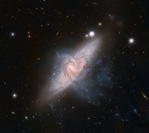 A space telescope view showing two large spiral galaxies, lined up one in front of the other. The foreground pinwheel-shaped galaxy is seen nearly face-on, with dark, swirling lanes of interstellar dust silhouetted against the bright glow of a larger background galaxy. The background galaxy is tilted both away from the viewer and roughly 45 degrees down to the left.