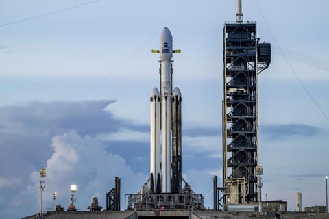 A white SpaceX Falcon Heavy rocket stands vertical on the black launch pad with a predawn sky in the background.