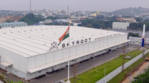 Aerial view of a large, low-slung warehouse-like building with a rocket standing outside it
