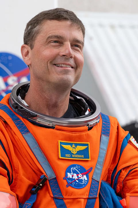 Reid Wiseman looks toward supporters, smiling, wearing an orange spacesuit with his name and a NASA meatball patch.