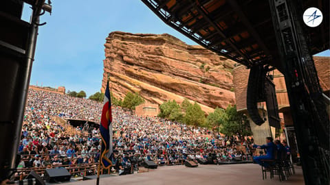 four people in blue flight suits stand on a stage in an amphitheater surrounded by red stone cliffs