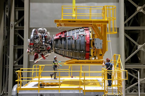 o In this image from Sept. 8, 2023, two technicians at NASA’s Michoud Assembly Facility in New Orleans stand around one of the four RS-25 engines on the core stage of the agency’s SLS (Space Launch System) rocket that would fly the Artemis II mission. The RS-25 engine test looks like a bell-shaped nozzle attached to a network of pipes. The engine is being held by the horizontal engine installer so it looks like it’s lying on its side. The RS-25 engine is about the size of a large pickup truck. T