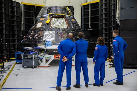 Four astronauts, wearing blue flight suits with their backs to the camera, look at the Orion spacecraft during the construction and testing process.
