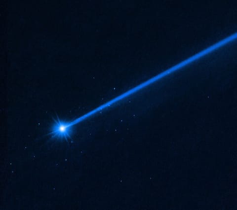 Against a black background, the bright white object at lower left is the asteroid Dimorphos. It has a bluish dust tail extending diagonally to the upper right. A cluster of blue dots surrounds the asteroid.