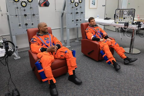 From left, NASA astronauts Victor Glover, Artemis II pilot and Reid Wiseman, Artemis II commander, are seated in chairs wearing orange jumpsuits. Behind them are two large gray panels with dials. The two astronauts are listening attentively and they prepare to conduct leak checks on his spacesuit inside the crew suit-up room in the Neil A. Armstrong Operations and Checkout Building at the agency’s Kennedy Space Center in Florida on Wednesday, April 1, 2026, ahead of the Artemis II test flight.