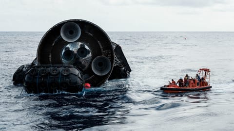 the aft end of a rocket with 3 small and 3 large engines floats in the ocean next to an orange raft with people aboard.