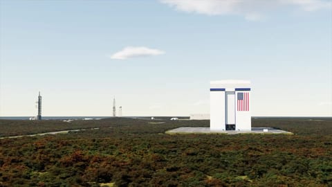 A square building stands on a flat landscape with a pair of rocket launch towers in the background.
