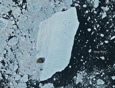 A detailed satellite view shows the iceberg wedged against the small, brown island, with sea ice packed densely to its left and more sparsely to its right.