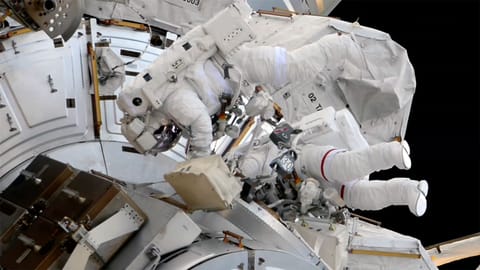 two astronauts in white spacesuits are seen outside a space station during a spacewalk