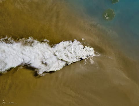 A white volcanic plume streams left over a tan ash cloud that fills most of the image. A small green island and blue water are visible in the upper right.