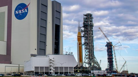 A bit orange rocket rolls out of a giant white building, with an extra launch tower in the background.
