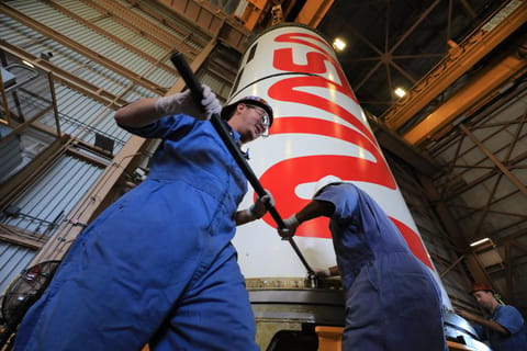Two workers in blue coveralls use long tools to secure hardware at the base of a large white rocket booster segment marked with a bold red NASA logo inside a high‑bay industrial facility. The camera angle is low, emphasizing the height of the booster above them, with structural beams and lighting visible overhead.