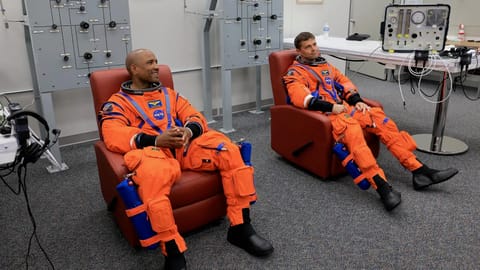 two male astronauts in orange flight suits sit in reddish recliners inside a white-walled room