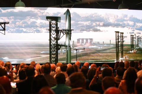 A crowd watches an animation showing multiple rockets launching from pads built over marshland