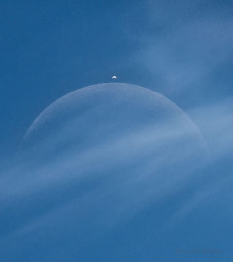 A light brown dust plume, dense at its center and more diffuse toward its edges, extends horizontally across an orange-brown and green landscape. Scattered white clouds cover some of the scene.