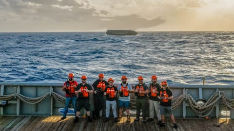 eight people stand on the deck of a ship at sea. behind them in the distance, a large black cylinder floats on top of the waves
