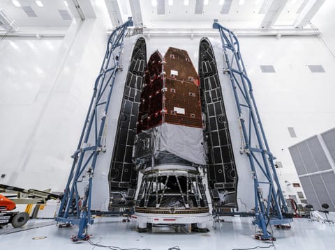 Full-height view of the Roman Space Telescope inside the tall payload fairing. Blue support structures stand on each side in the cleanroom environment.