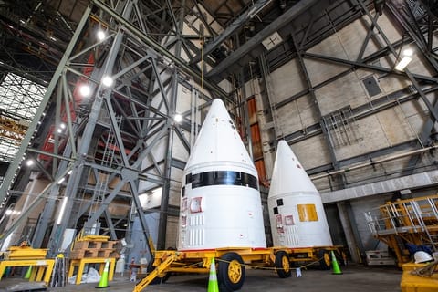Two large white rocket-shaped payload fairings sit on yellow transport carts inside a tall industrial facility. The building’s interior is filled with steel beams, platforms, and bright overhead lights.