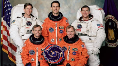Five astronauts, two in white spacewalking suits and three in orange launch suits, post for an STS-69 NASA shuttle crew portrait.