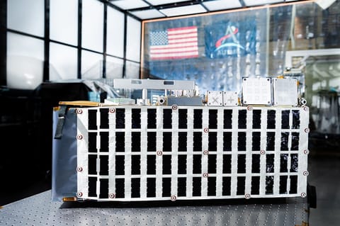 Rectangular box-shaped device resting on a metal table. The front face is covered with a grid of many small black rectangular panels bordered in white. Metal components, brackets, and small box units are mounted along the top. The background shows a large windowed wall with a blurred American flag and an Artemis flag visible behind the device.