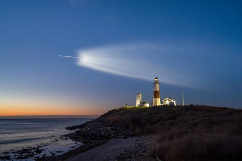 A rocket in launch over the ocean with a lighthouse in the foreground.
