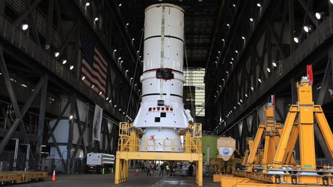The bottom segment of a white solid rocket booster stands on a yellow support inside a giant rocket hangar.