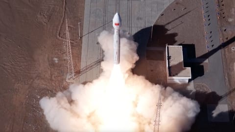 aerial drone photo of a white rocket launching from a desert pad