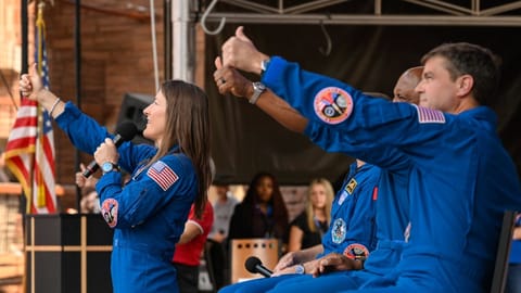 four people in blue flight suits stand on a stage in an amphitheater surrounded by red stone cliffs