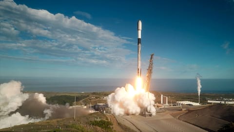 A white, black and gray-ish (soot-covered) rocket lifts off into a mostly-blue sky from a sea-side launch pad.