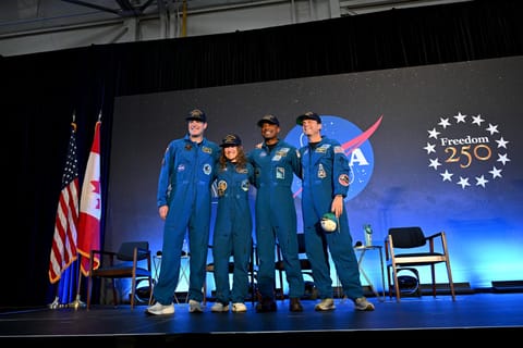 The four Artemis II crew members stand side-by-side and smiling in their blue flight suits on a stage at NASA Johnson. Bel