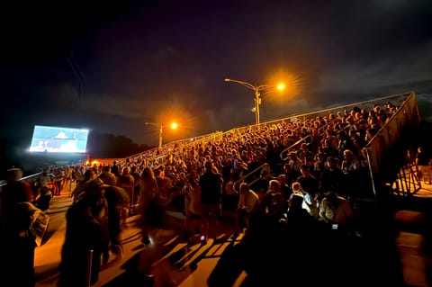 A large crowd gathered along a waterfront at night to watch a launch, with a screen and lights visible in the distance.