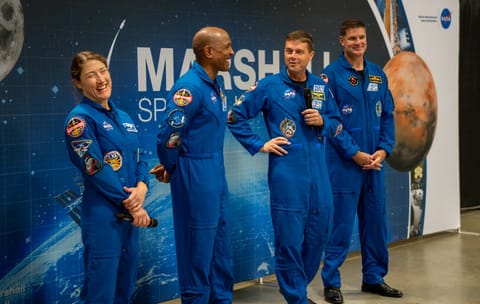 The Artemis II crew smile and laugh. They are wearing blue jumpsuits with patches on them. Christina Koch, left, and Reid Wiseman, second from right, are holding microphones.