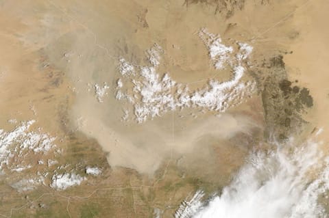 A light brown dust plume, dense at its center and more diffuse toward its edges, extends horizontally across an orange-brown and green landscape. Scattered white clouds cover some of the scene.