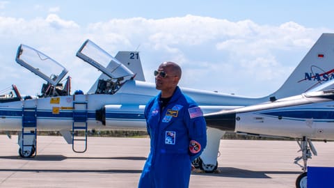 A man in a blue jumpsuit stands in front of giant a trio of NASA jets.