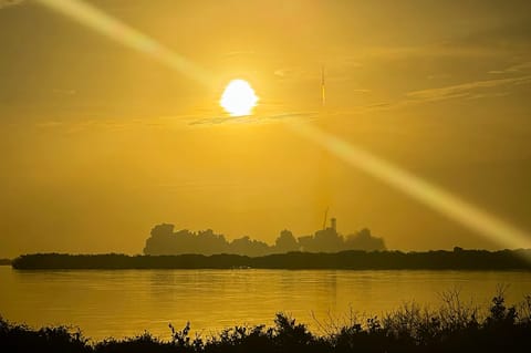 A rocket standing on its launch pad near the horizon at sunrise, seen across water with a bright sun overhead.