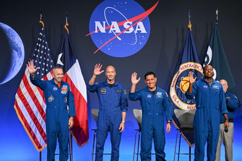 Four astronauts are pictured wearing their blue flight suits, smiling and waving. They are all on-stage with four chairs behind them. Behind the chairs is the NASA meatball, with American and Texas flags to the left and NASA and ESA (European Space Agency) flags to the right.