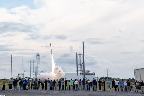 In the foreground, a line of people stand at a safe distance away from a sounding rocket launching in the background. The rocket is just off the pad with a plume of fire and smoke underneath.