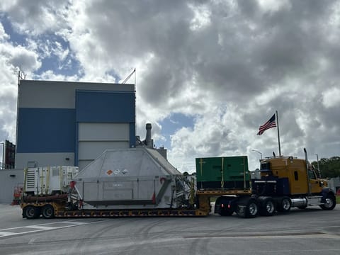 A large truck with a yellow cab hauls a wide, metal, container-like structure on a low trailer, with additional equipment secured behind it. An American flag flies from the truck as it sits on a paved area beside a tall industrial building. Thick, backlit clouds fill the sky above at NASA's Kennedy Space Center in Florida on Tuesday, April 28, 2026. Photo credit: NASA/Tiffany Fairley
