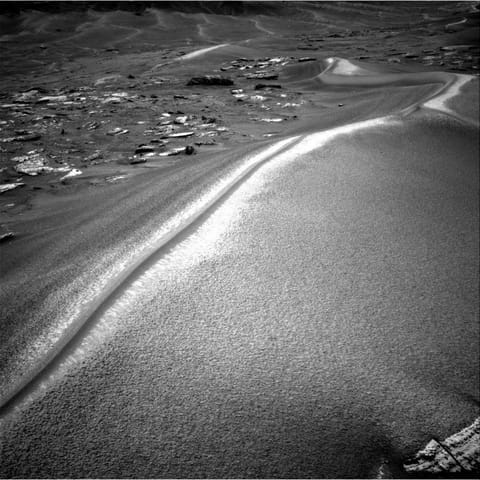 A black-and-white photograph from the Mars Curiosity rover showing a large, sweeping sand dune with a sharp, brightly sunlit crest. To the left are scattered flat rocks, and more rolling dunes stretch into the distant background.
