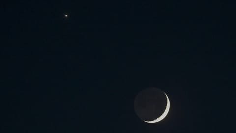 A thin crescent moon shines in a black sky with Venus appearing as a bright star-like object to its upper left.