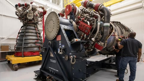 Two large rocket engines, one standing, one horizontal, rest on support platforms inside a white industrial setting.